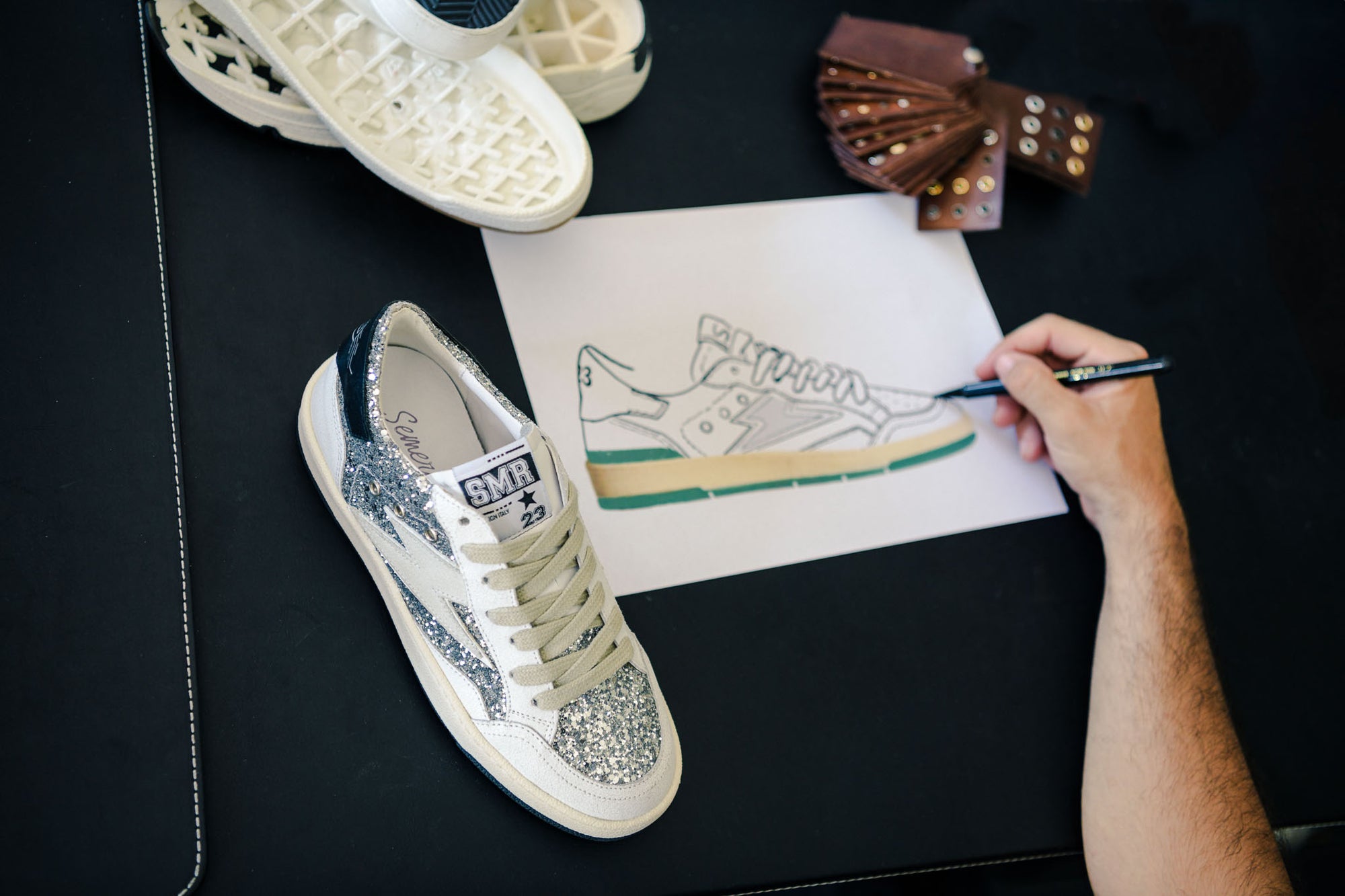 Designer de chaussures dessinant un nouveau modèle de baskets Semerdjian SMR à paillettes sur une table de travail.